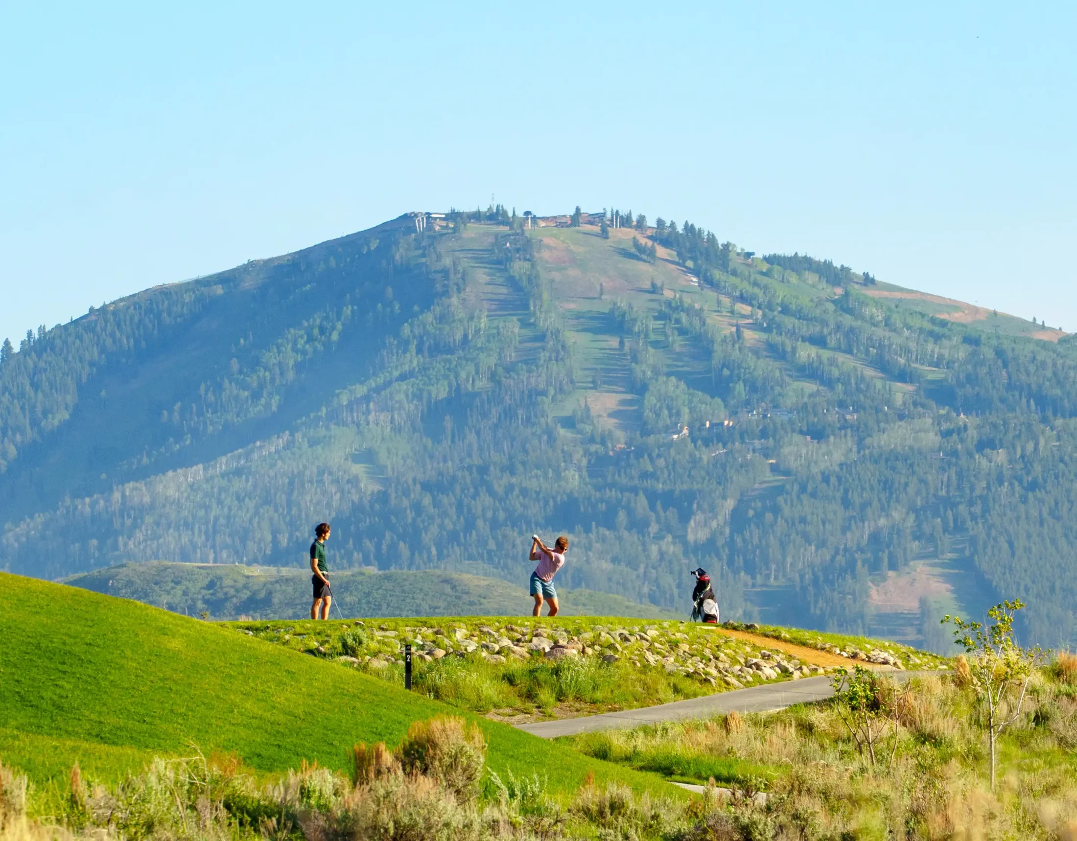 Two golfers standing on a ridge teeing off with a mountain in the background.